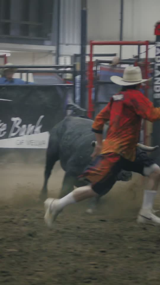 A bullfighter in orange sprinting ahead of a charging black bull